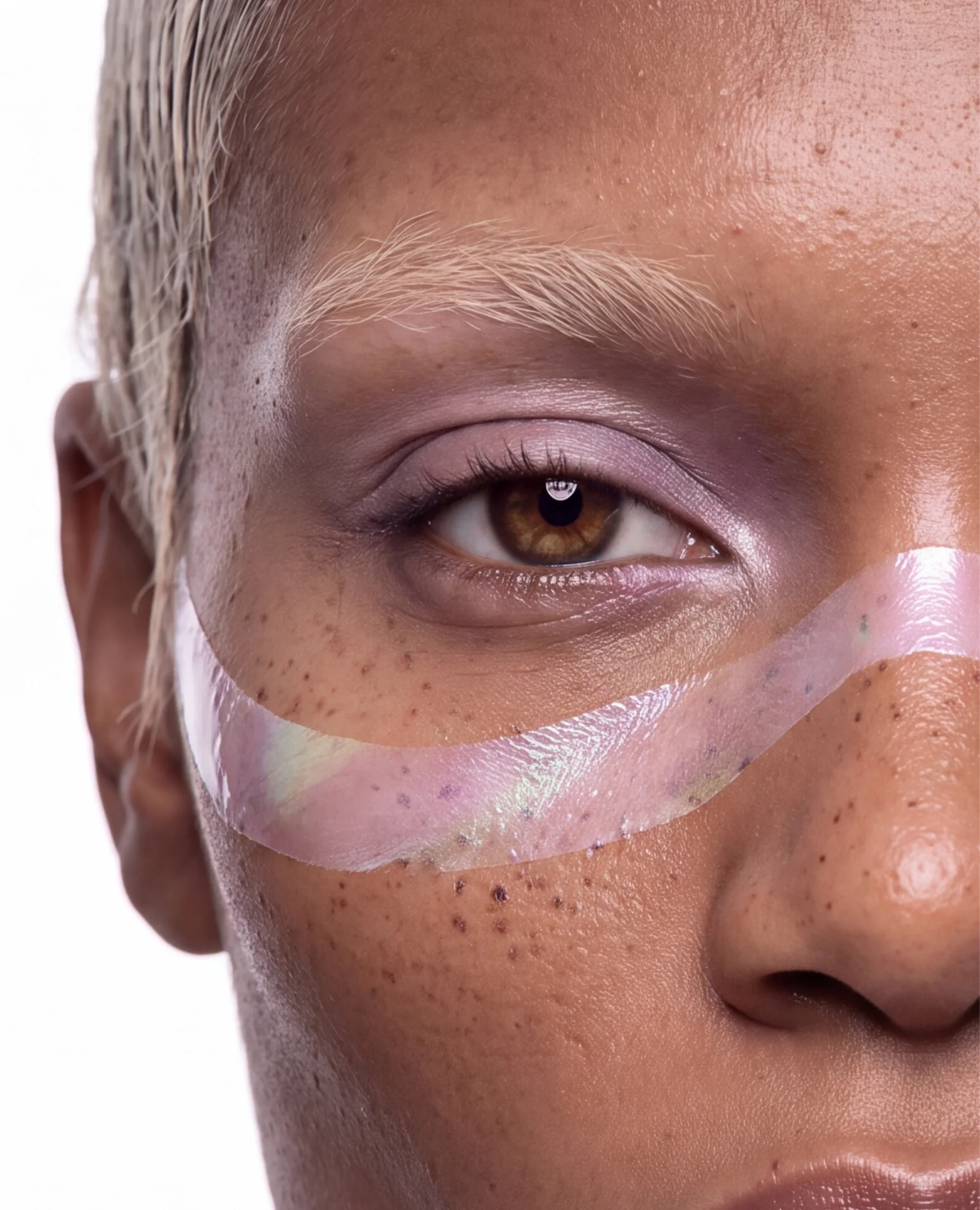 Brown eye with lilac shadow and an iridescent ribbon across freckled skin.