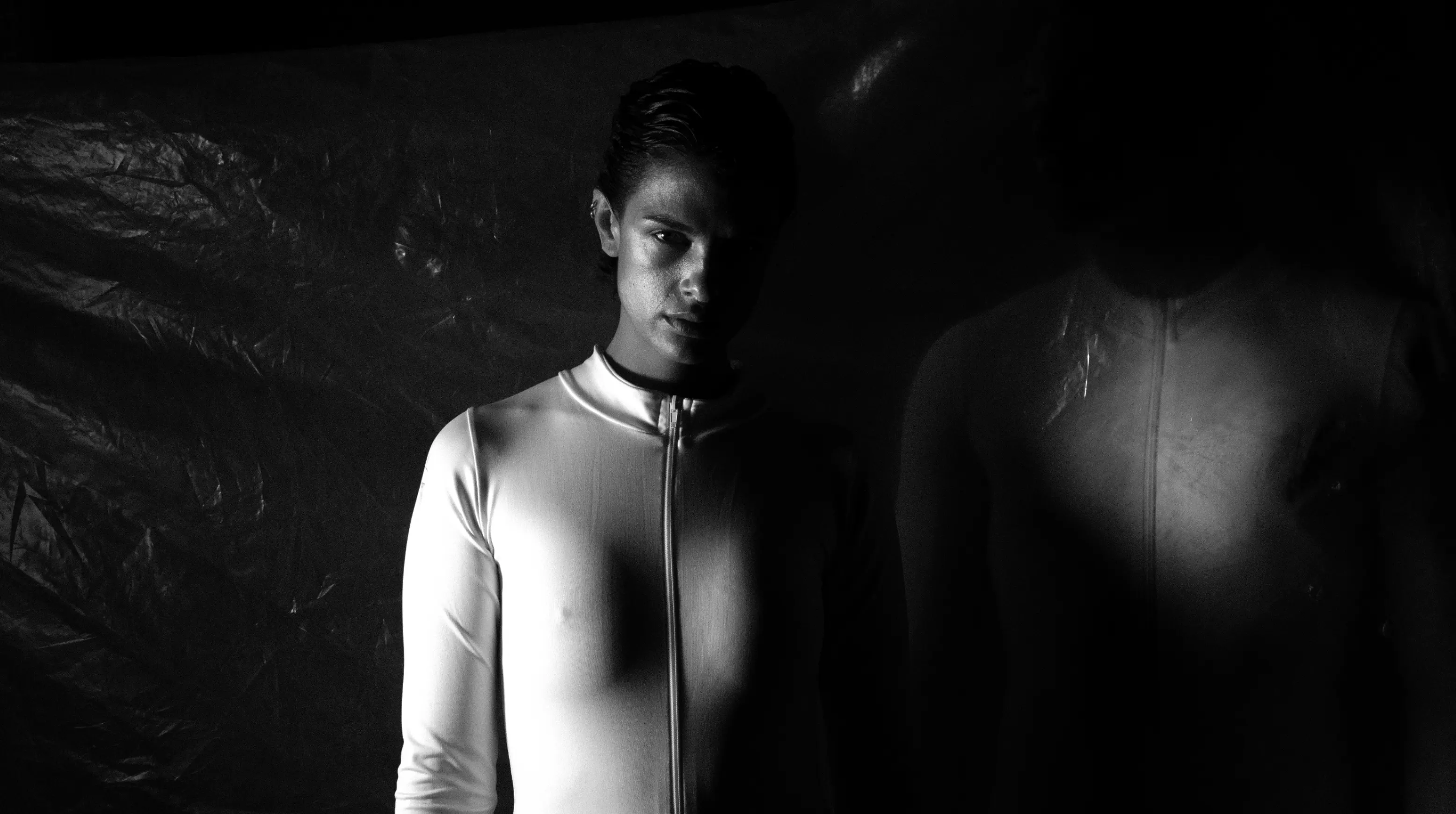 A man in a white suit against crumpled black plastic, a figure in shadow beside him.