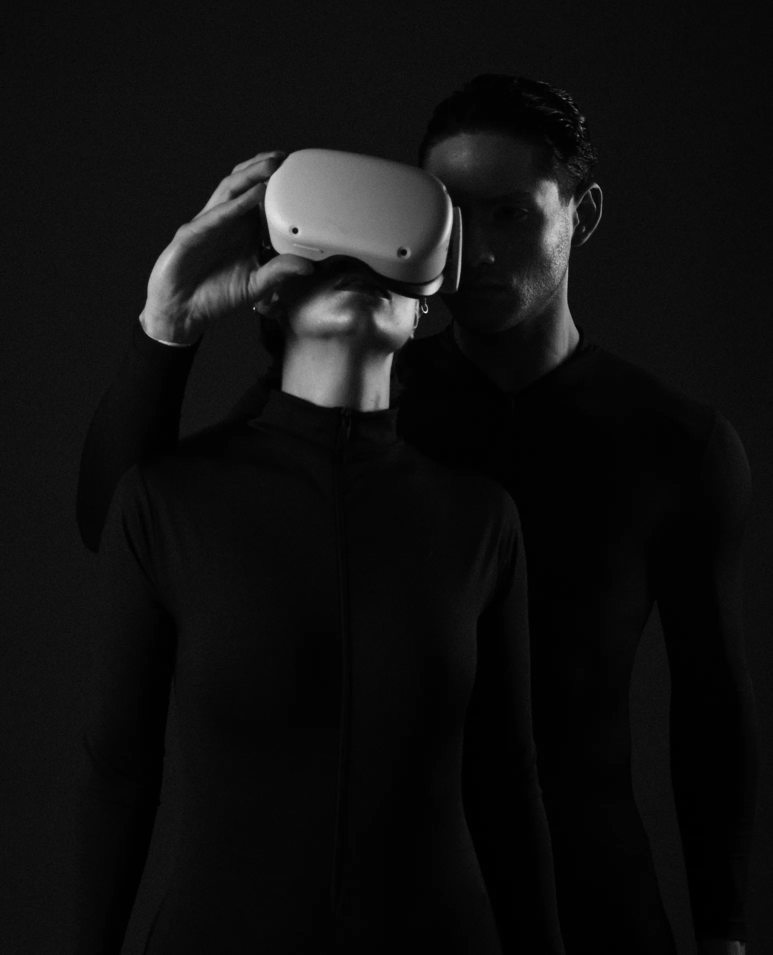 A woman lifting a VR headset in the dark as a man leans in behind her.